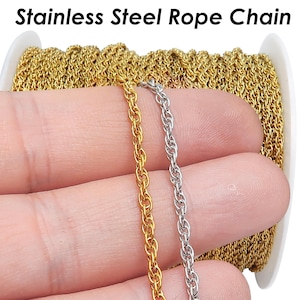 Puede incluir: Cadena de cuerda de acero inoxidable en oro y plata. La imagen muestra dos hebras de cadena, una dorada y otra plateada, sostenidas en una mano. Las cadenas están junto a un carrete de cadena dorada. El texto "Stainless Steel Rope Chain" está en la parte superior.