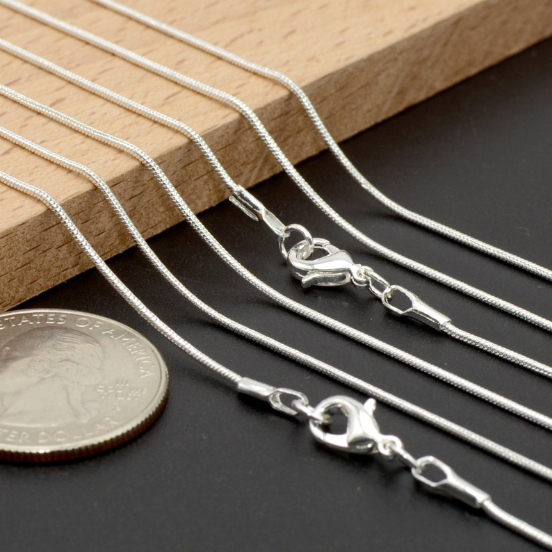 May include: A set of six silver snake chains of varying lengths. The chains are laid out on a wooden surface and a US quarter is included for scale.