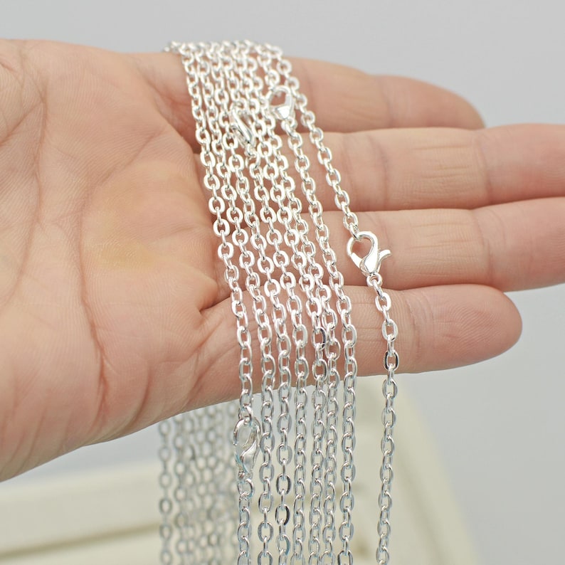 May include: A close-up of a silver chain necklace with a lobster clasp. The chain is shown in multiple lengths, with the longest chain being held in a hand.