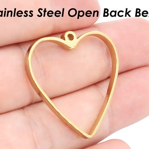 Könnte beinhalten: Vergoldeter, offener Herz-förmiger Bezel-Anhänger aus Edelstahl. Der Anhänger hat eine glatte, polierte Oberfläche und eine Öse oben zum Anbringen einer Kette oder Schnur.