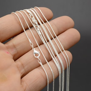 May include: A set of silver snake chains of varying lengths, displayed on a hand. The chains are delicate and have a smooth, polished finish.