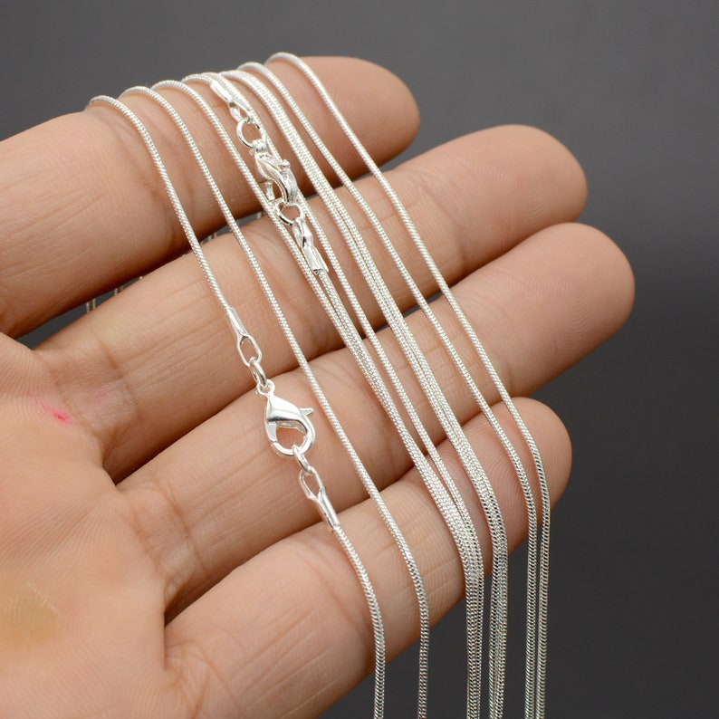 May include: A set of silver snake chains of varying lengths, displayed on a hand. The chains are delicate and have a smooth, polished finish.