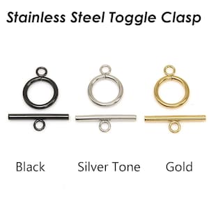 Könnte beinhalten: Edelstahl-Knebelverschlüsse in Schwarz, Silber und Gold. Jeder Verschluss besteht aus einem Ring und einem Stab mit einer kleinen Schlaufe. Der Text "Stainless Steel Toggle Clasp" steht oben, mit den Farben darunter.