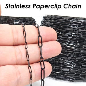 Könnte beinhalten: Schwarze Edelstahl-Büroklammerkette. Das Bild zeigt eine Nahaufnahme der Kette, wobei ein Teil in einer Hand gehalten wird. Die Kette besteht aus kleinen, länglichen Gliedern. Der Text "Stainless Paperclip Chain" ist oben sichtbar.
