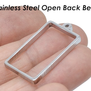 May include: A silver open back bezel pendant with a rectangular shape. The pendant has a loop at the top for attaching a chain or cord.