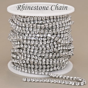 Puede incluir: Una cadena de pedrería plateada enrollada en una bobina blanca. Una pequeña longitud de la cadena está desenrollada en la parte inferior. El texto "Rhinestone Chain" está impreso en la parte superior de la bobina. Ideal para bisutería.