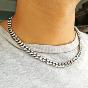 Op de afbeelding: Een zilverkleurige gourmette ketting. De ketting is gemaakt van in elkaar grijpende schakels en wordt om de nek gedragen. De ketting is dik en heeft een glanzende afwerking. De persoon draagt een grijs T-shirt.