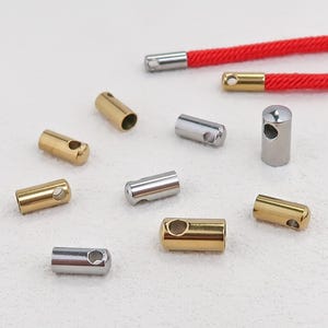 Puede incluir: Una colección de pequeños terminales cilíndricos de metal en tonos oro y plata, cada uno con un orificio lateral para su fijación. Dos de los terminales están unidos a cordones trenzados de color rojo brillante, los demás están dispersos.