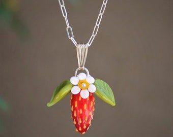 May include: A handcrafted necklace featuring a red strawberry pendant with white and yellow accents, green leaves, and a white flower. The pendant hangs from a silver chain, creating a charming and unique piece of jewelry.