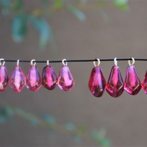 May include: Ten pink glass teardrop-shaped charms with silver jump rings hanging from a black string.