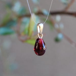 May include: A gold pendant with a teardrop-shaped, deep red gemstone. The pendant hangs from a thin, clear cord. The background is blurred, with hints of green foliage.