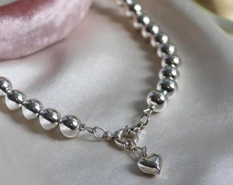May include: A silver-toned beaded necklace featuring a series of polished, spherical beads connected by metal links. The necklace includes a circular spring-ring clasp at the front, from which a small, smooth, heart-shaped charm is suspended.