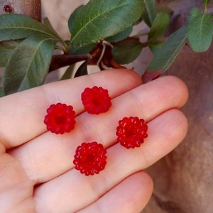 Pode incluir: Quatro pequenas contas de vidro redondas e de cor vermelho brilhante com uma superfície texturizada e rugosa que imita a aparência de framboesas. Cada conta tem um orifício central e estão dispostas em grupo, exibindo a sua cor vermelha translúcida.