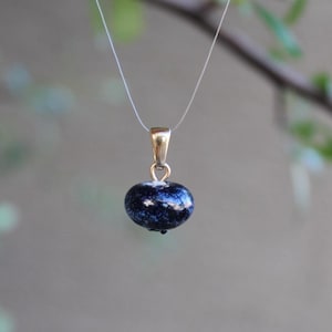 May include: A gold-colored pendant with a dark blue, speckled bead. The bead is suspended from a thin, clear cord. The pendant is set against a blurred background of green leaves and a neutral gray backdrop.