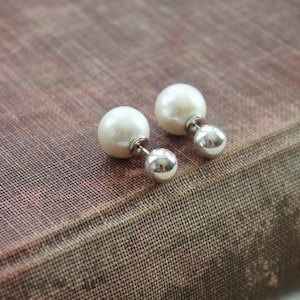 Può includere: Un paio di orecchini a doppio lato con una grande perla sferica color bianco sporco e una sfera più piccola in metallo lucido color argento. Le due sfere sono collegate da un perno metallico dritto, creando un design bilanciato.