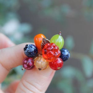 May include: A cluster ring featuring a variety of small, realistic fruit-shaped beads including a bright red raspberry, a green gooseberry, an orange, a translucent beige berry, and several dark blue and red speckled berries with a small metal leaf.