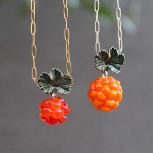 May include: Two necklaces featuring pendants shaped like cloudberries. One pendant is bright orange and the other is a deep red. Each berry is topped with a textured, dark green metallic leaf. The necklaces hang from gold and silver link chains.
