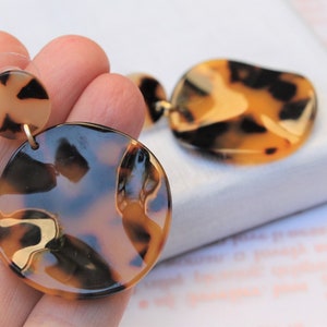 May include: Tortoiseshell-patterned earrings. The earrings feature a small, round stud and a larger, irregular-shaped pendant. The color palette includes shades of brown, tan, and black, with a glossy finish. The earrings are designed for pierced ears.
