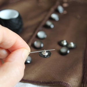 May include: A small, round, dark gray button with a metallic shank is held by a silver sewing needle. Several identical buttons are arranged in a line on a piece of brown, semi-sheer fabric, with a black spool of thread visible in the background.