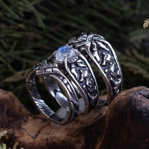 Puede incluir: Tres anillos de plata con diseños intrincados, uno con una piedra de luna. Los anillos están apilados sobre un trozo de madera.