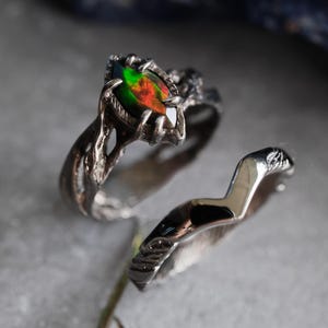 May include: Two silver rings. One ring features a marquise-cut gemstone with green, red, and orange hues, set in a textured, organic-style band. The second ring has a smooth, curved design with a textured band.