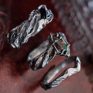 Może przedstawiać: Trzy srebrne pierścionki o skomplikowanych, organicznych wzorach. Jeden pierścionek ma ciemny kamień szlachetny o czerwonych i zielonych odcieniach. Pierścionki są prezentowane na ciemnej, teksturowanej powierzchni, pokazując ich kunszt.