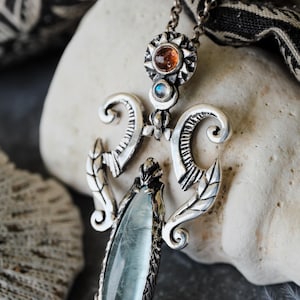 Può includere: Una collana con un ciondolo in argento decorato con una grande pietra di acquamarina blu chiaro incastonata in un'impostazione in argento dettagliata e elaborata. Il ciondolo presenta un design a forma di sole con una piccola pietra arancione nella parte superiore.