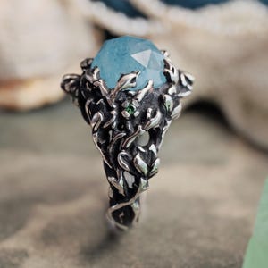 May include: A silver ring featuring a large, light blue gemstone with multiple facets. The ring's band and setting are detailed with a vine and leaf pattern, and a small green stone is set into the design.