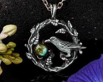 May include: A silver-toned pendant necklace featuring a detailed raven design within a circular frame of leaves. The raven is perched near a round, iridescent stone. The necklace hangs on a silver chain, with a dark background and floral accents.