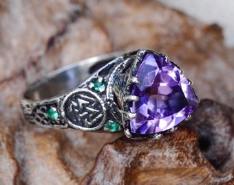 May include: A silver ring featuring a large purple amethyst gemstone and two smaller green gemstones. The ring has an intricate design with a symbol that looks like a triquetra.