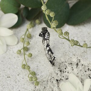 Peut inclure: Une bague argent&eacute;e avec un motif de feuilles d&eacute;taill&eacute;. La bague est incurv&eacute;e et pr&eacute;sente des motifs complexes, avec un petit d&eacute;tail floral. Le fond comprend des fleurs blanches et du feuillage vert, cr&eacute;ant une esth&eacute;tique naturelle.