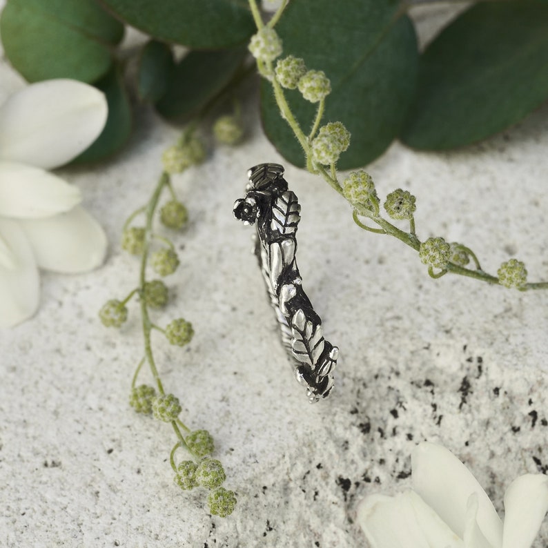 Peut inclure: Une bague argent&eacute;e avec un motif de feuilles d&eacute;taill&eacute;. La bague est incurv&eacute;e et pr&eacute;sente des motifs complexes, avec un petit d&eacute;tail floral. Le fond comprend des fleurs blanches et du feuillage vert, cr&eacute;ant une esth&eacute;tique naturelle.