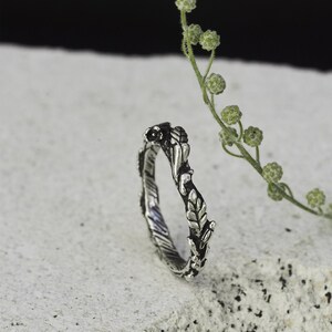 Peut inclure: Une bague argent&eacute;e orn&eacute;e d'un motif de feuilles et de vigne. La bague pr&eacute;sente des feuilles et des vignes d&eacute;taill&eacute;es qui entourent l'anneau. La bague est pos&eacute;e sur un fond neutre avec une brindille de verdure.