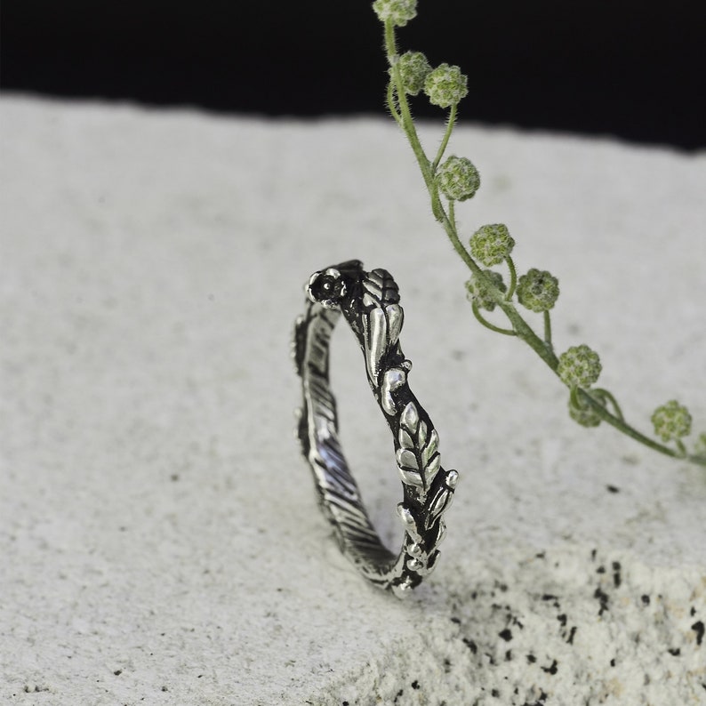 Peut inclure: Une bague argent&eacute;e orn&eacute;e d'un motif de feuilles et de vigne. La bague pr&eacute;sente des feuilles et des vignes d&eacute;taill&eacute;es qui entourent l'anneau. La bague est pos&eacute;e sur un fond neutre avec une brindille de verdure.