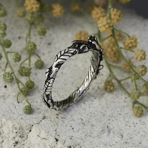 Peut inclure: Une bague argent&eacute;e orn&eacute;e d'un motif inspir&eacute; de la nature. La bague pr&eacute;sente un motif d&eacute;taill&eacute; de feuilles et de vignes, cr&eacute;ant un aspect textur&eacute; et organique. La bague est mise en valeur sur un fond de verdure.