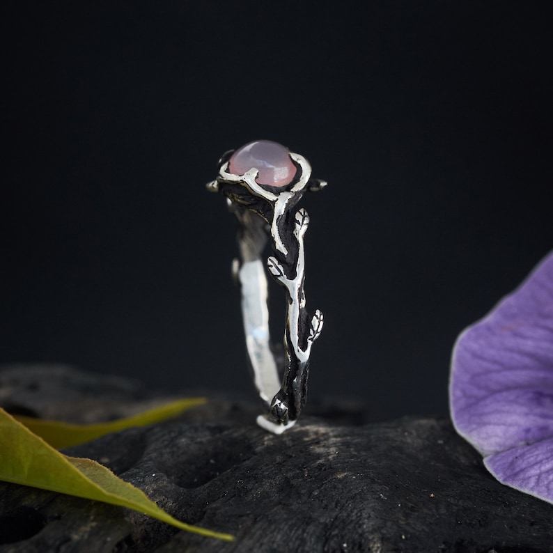 May include: A silver ring with a pink gemstone set in a branch-like design. The ring is oxidized for a darker, more rustic look.