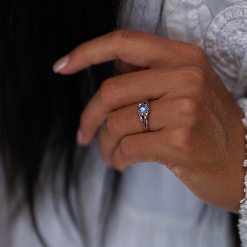 Puede incluir: Un delicado anillo con una piedra de luna azul engastada en una banda de oro rosa. El anillo est&aacute; dise&ntilde;ado de forma intrincada con un patr&oacute;n de espiral.