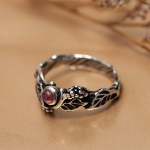 Puede incluir: Un anillo de plata con una piedra preciosa rosa engastada en un diseño floral. El anillo presenta hojas y bayas.