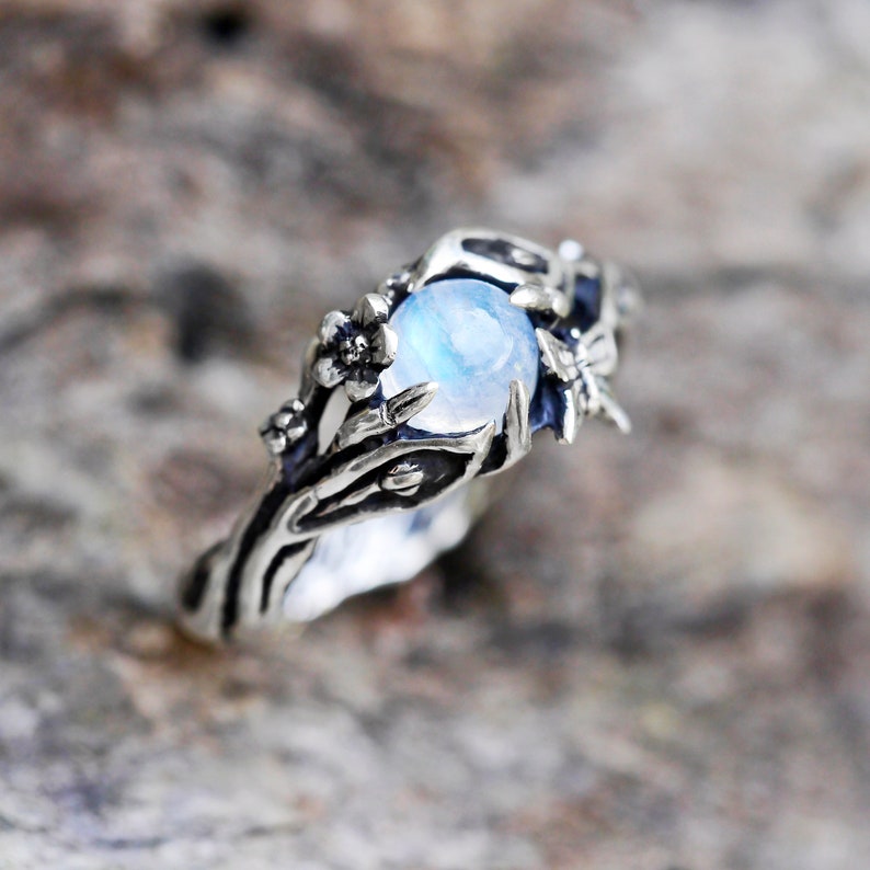 May include: A silver ring with a moonstone set in a floral design. The ring features delicate branches and flowers surrounding the gemstone.