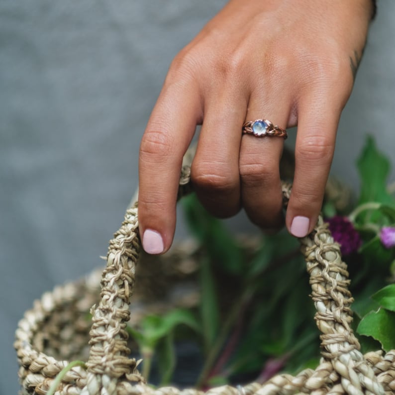 Puede incluir: Una mano con un anillo con una piedra preciosa azul claro engastada en una banda de oro rosa. La mano sostiene una cesta tejida llena de plantas verdes y flores moradas.