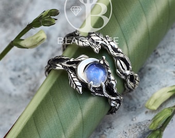 Puede incluir: Un par de anillos de plata con una piedra de luna engastada en un diseño de media luna. Los anillos están entrelazados con ramas y hojas.