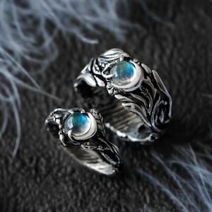 May include: Two silver rings with intricate floral and leaf designs. Each ring features a large, round, blue-green gemstone. The rings are displayed on a dark, textured surface with soft, white feather accents.