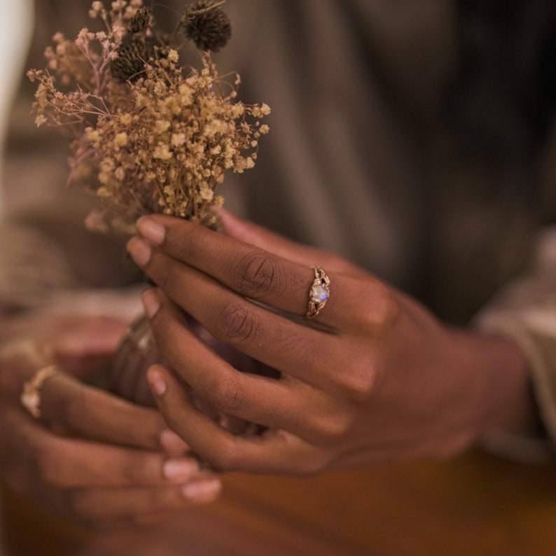 Puede incluir: Primer plano de una mano con un anillo de oro con una piedra preciosa transparente. El anillo est&aacute; en el dedo medio de la mano. La mano sostiene un ramo de flores secas.