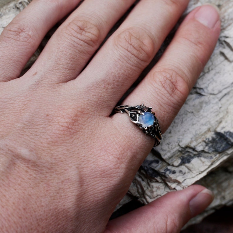 May include: A silver ring with a moonstone set in a twisted vine design. The ring is on a hand, with the background out of focus.