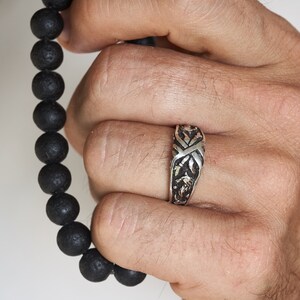 May include: A silver ring with an intricate design. The ring is worn on the middle finger of a hand.
