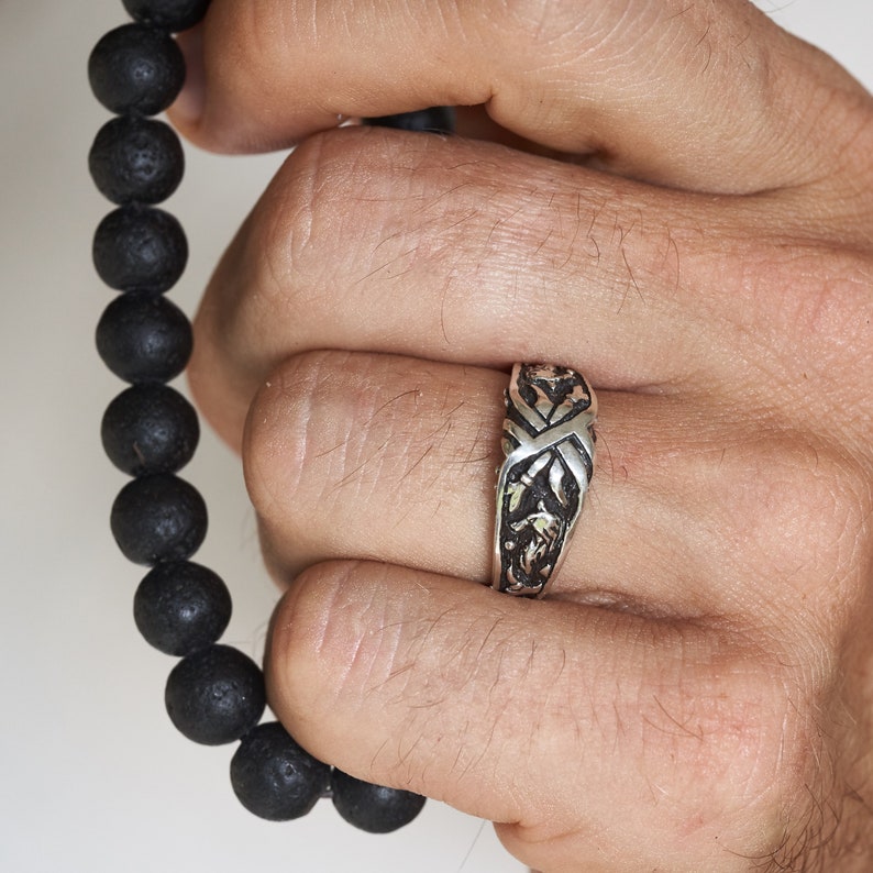 May include: A silver ring with an intricate design. The ring is worn on the middle finger of a hand.
