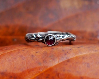 May include: A silver ring with a dark red gemstone set in a twig-like band.