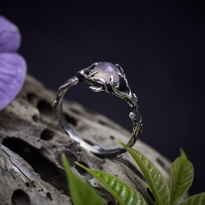 May include: A silver ring with a delicate branch design and a pale purple gemstone. The ring is set on a piece of driftwood with green leaves.