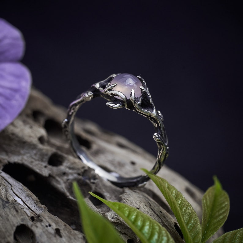 May include: A silver ring with a delicate branch design and a pale purple gemstone. The ring is set on a piece of driftwood with green leaves.