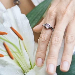 May include: A silver ring with a delicate floral design and a pale pink gemstone. The ring is worn on a finger against a backdrop of white flowers.